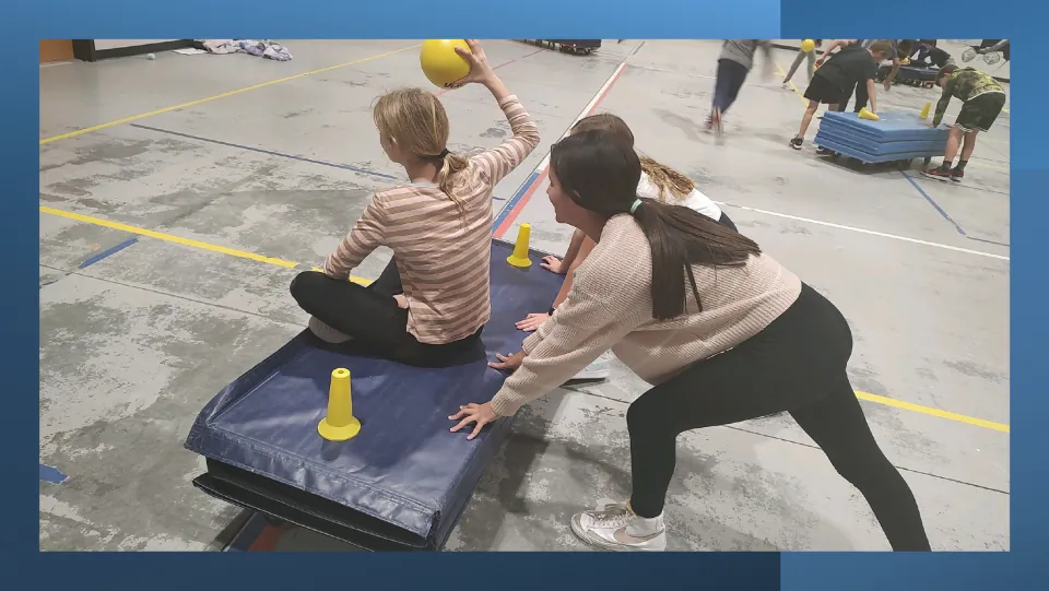Game in action: a seated bomber raises a yellow ball to throw while a teammate pushes the mat boat with yellow cones
