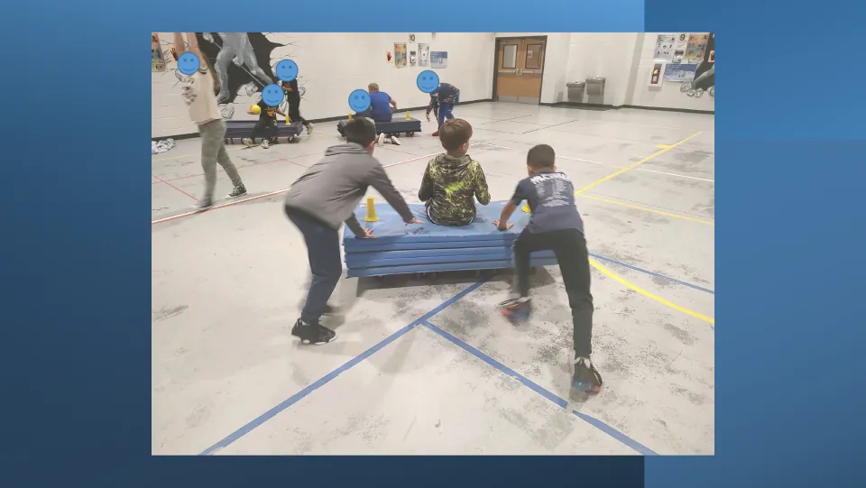 Two pushers drive a stacked-mat boat across the gym floor as a seated rider holds on, with other boats playing behind