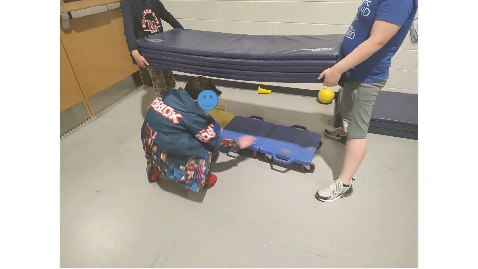 Boat building: a student lines up scooter boards while two others hold a folded gym mat ready to set on top
