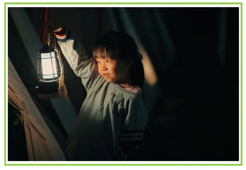 Camping writing and discussion prompt card 5: a child holding up a glowing lantern beside a tent opening